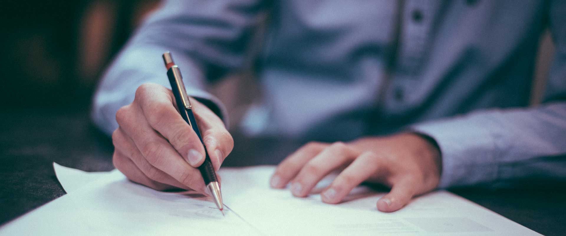 Man in blue-collared dress shirt signs a document with a black and gold pen